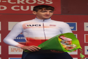Ein Mann steht triumphierend auf einem Podium, hält einen Blumenstrauß in der Hand und hat eine Tafel im Hintergrund.
