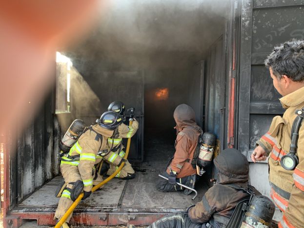 Feuerwehrleute in Helmen und Schutzausrüstung arbeiten von der Ladefläche eines raucherfüllten Trucks, wobei einer einen Schlauch hält, um ein Feuer zu löschen.