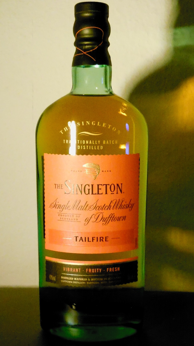 Eine tiefe Bernsteinflasche Singleton of Dufftown Tailfire Single Malt Scotch Whisky auf einem Tisch, mit einem schwarzen Etikett, das den Namen des Whiskys und ein Wappen mit einem Löwen und einer Burg zeigt.