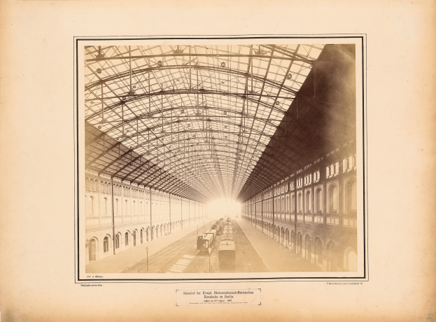 Schwarzes und weißes Foto eines Bahnhofs mit einem Zug auf den Schienen, umgeben von Gebäuden mit Fenstern und einem Dach, das von Metallpfosten gestützt wird, mit Text unten.