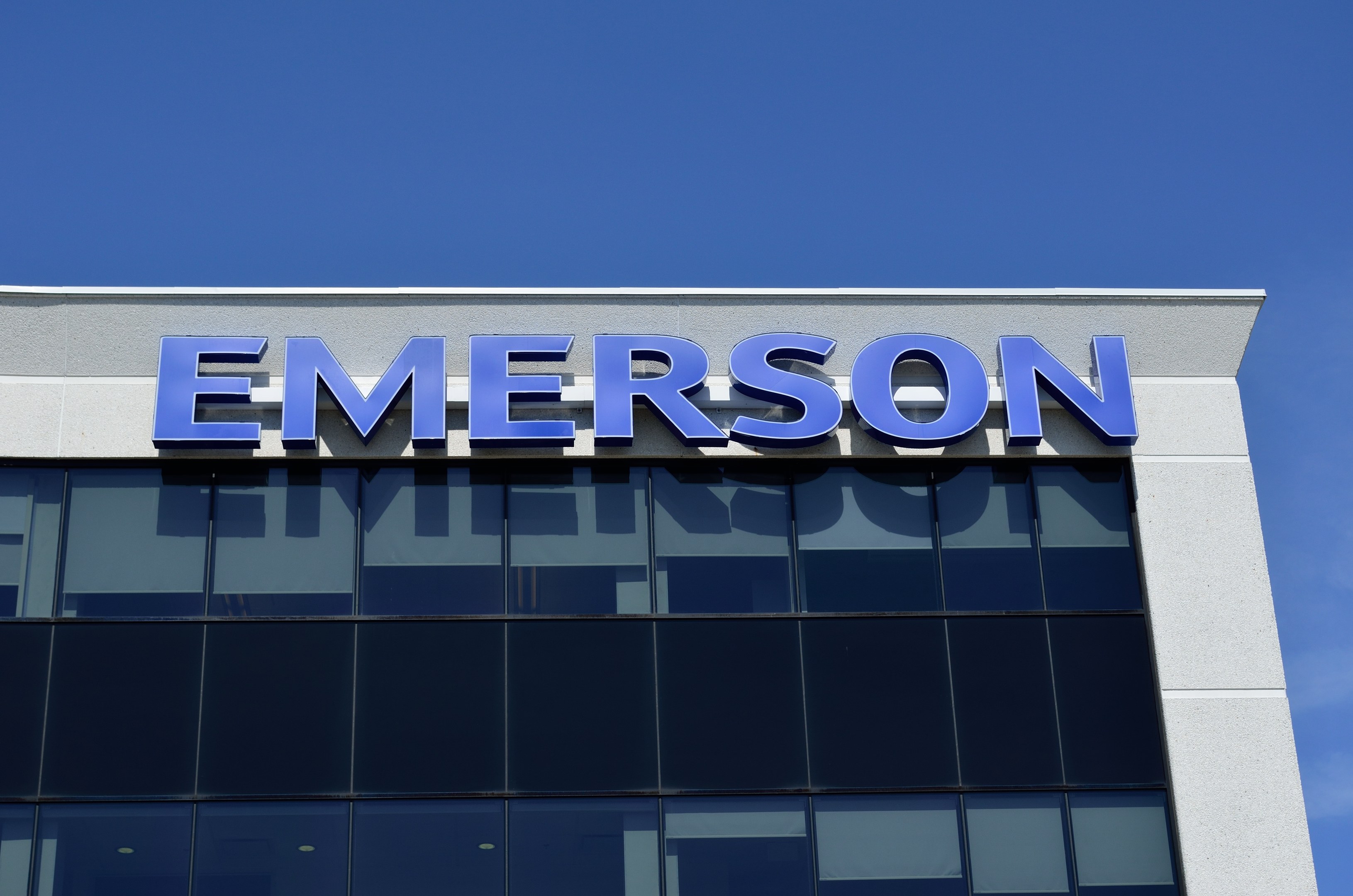 Das Emerson-Logo an der Seite eines Gebäudes mit dem Text "Emerson investiert 1,5 Milliarden US-Dollar in den USA" vor einem klaren blauen Himmel.