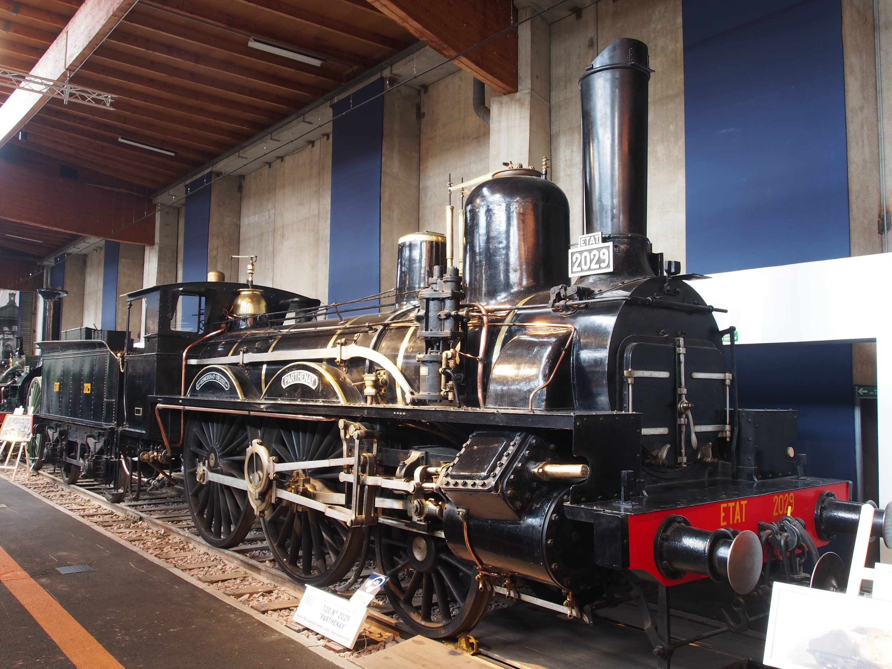 Eine große, alte Dampflokomotive in einem Museums Setting umgeben von Eisenbahnschienen, Informationsschildern und anderen Ausstellungsstücken, mit einer Wand und beleuchteter Decke im Hintergrund.