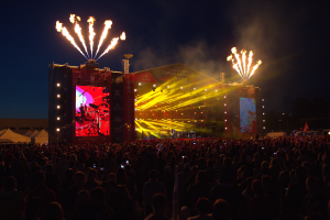 Eine große Menschenmenge versammelte sich auf einem Musikfestival, mit einer Bühne in der Mitte, auf der Musiker von hellen Scheinwerfern beleuchtet wurden und von Bannern umgeben waren, Zelte links und ein dunkler Himmel mit Feuerwerk im Hintergrund.