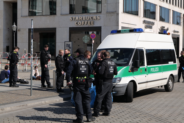 Eine Gruppe von Polizeibeamten steht vor einem Starbucks-Café, rechts ein Van und links einige Menschen; im Hintergrund ist ein Gebäude mit Fenstern, ein Schild, ein Laternenmast und ein Zaun zu sehen.