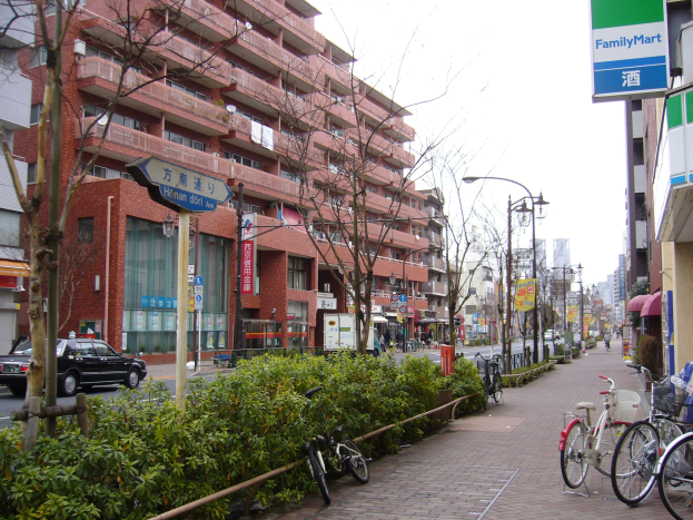 Eine Stadtstraße mit parkenden Fahrrädern, Gebäuden, Straßenlaternen, Schildern, fahrenden Fahrzeugen, Fußgängern auf dem Gehweg, Grünfläche und einem klaren blauen Himmel.