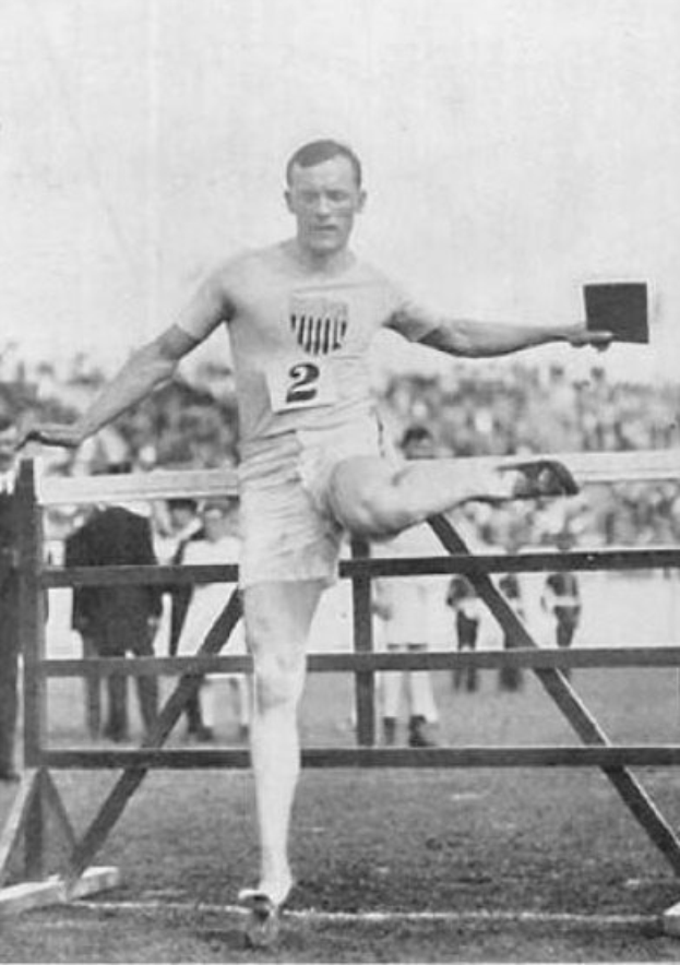Schwarzes und weisses Foto eines Mannes in der Luft, der über ein Hindernis springt, mit einer nummerierten Shorts und einem T-Shirt, mit Zuschauern im Hintergrund und Himmel darüber.