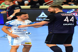 Zwei Männer geben sich die Hand auf einem Futsal-Platz vor Publikum, mit Zuschauern auf Stühlen und Geländern sowie einem Anzeigebildschirm im Hintergrund, wobei einer der Männer Schuhe trägt.