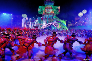 Eine Gruppe von Menschen in bunten Kostümen tanzt auf einer Bühne vor einem sitzenden Publikum, mit Lichtern, Pfählen, Luftballons und einem Bildschirm, der eine Person im Hintergrund zeigt, unter einem sichtbaren Himmel, bei der Rio-Karneval 2014.