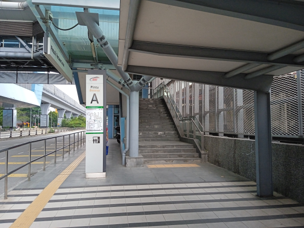 Ein Bahnsteig mit Treppe, eine Straße mit Geländern auf der linken Seite und ein Hintergrund mit Bäumen, Pfählen, Gebäuden und einer Brücke, mit Rohren und einer Tafel oben auf dem Bahnsteig.