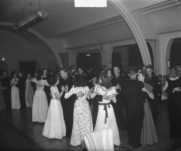 Schwarzes und weißes Foto von Menschen, die in einem Ballsaal tanzen, mit Deckenlichtern und Stühlen im Hintergrund.