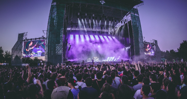 Eine große Menschenmenge auf einem Musikfestival, mit einer Bühne in der Mitte, auf der Musiker Instrumente spielen, beleuchtet von Scheinwerfern und umgeben von Bildschirmen, mit einem Gebäude, Bäumen und einem klaren blauen Himmel im Hintergrund.