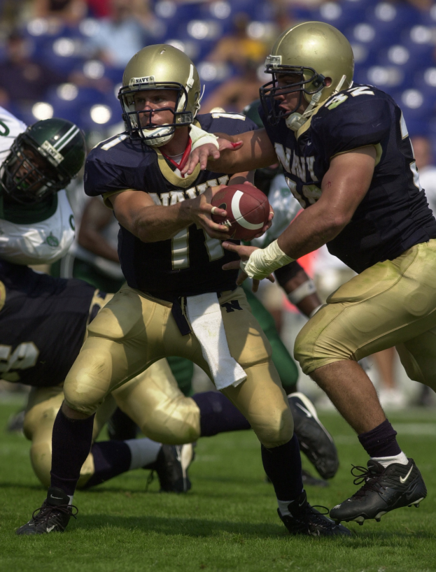 Ein Fußballspieler in einem Sportdress und Helm rennt mit dem Ball, umgeben von anderen Spielern in ähnlicher Kleidung, mit einem unscharfen Hintergrund.