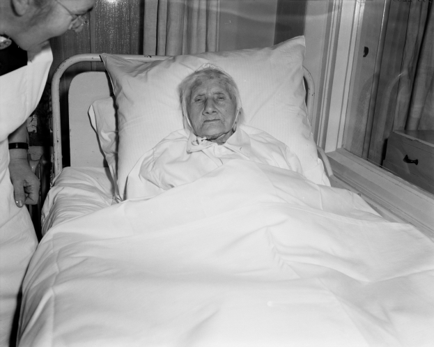 Ein älteres Frau liegt in einem Krankenhausbett mit einer weißen Decke und Kissen, während eine Krankenschwester neben ihr steht, mit einem Fenster mit Vorhängen im Hintergrund.